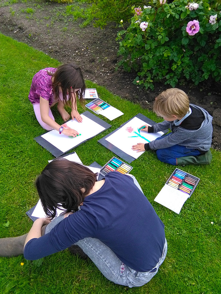 3 enfants qui dessinent assis par terre dans le jardin